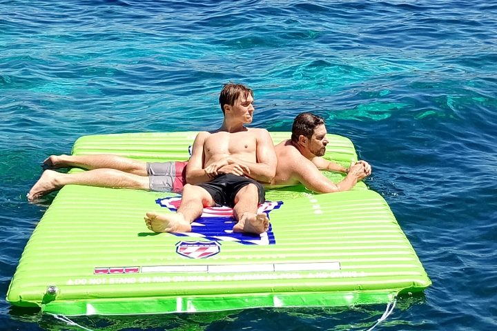 Two men relaxing on a green inflatable raft in the ocean with a distant island background.