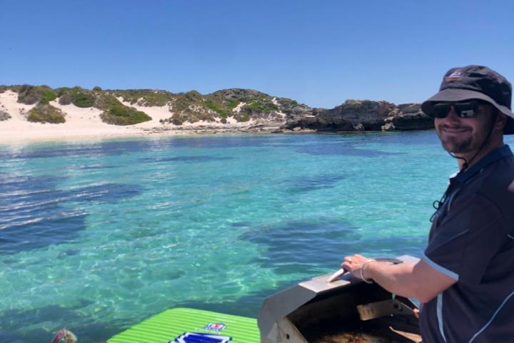 Person grilling on a boat, another on colorful float in clear water near a sandy beach.