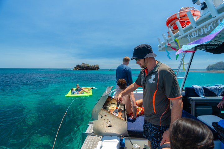 Man grilling on boat with people relaxing in ocean on a sunny day.