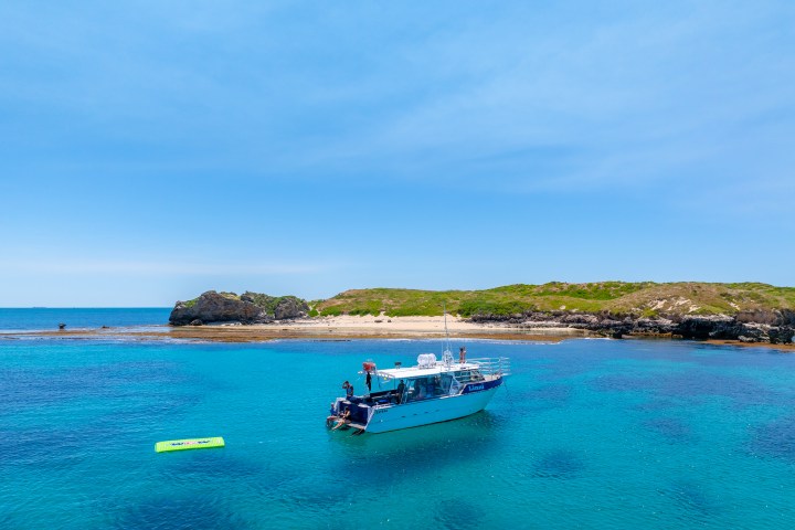 Boat anchored in clear blue sea near a rocky island with green vegetation under a bright sky.