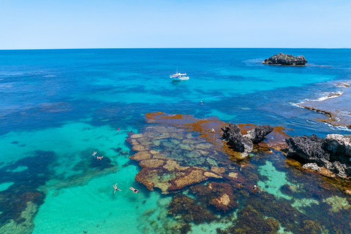 Aerial view of people snorkeling near rocks in clear blue ocean with distant boat.