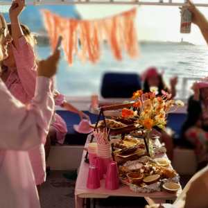 Group of women celebrating on a boat with food and drinks, dressed in pink and enjoying sunny weather.