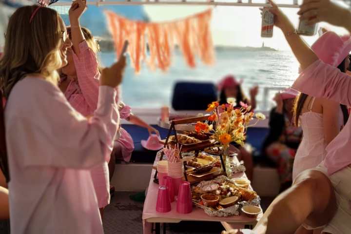 Group of women celebrating on a boat with food and drinks, dressed in pink and enjoying sunny weather.