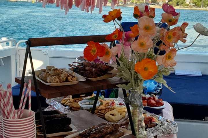Buffet table on a boat with snacks, flowers, pink cups, and a pink ribbon decoration in the background.