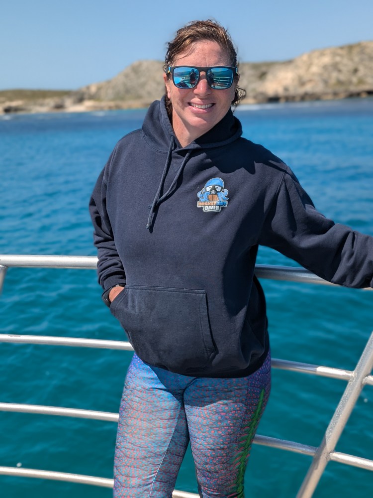 Person in hoodie and sunglasses standing on a boat with ocean and rocky coastline in the background.