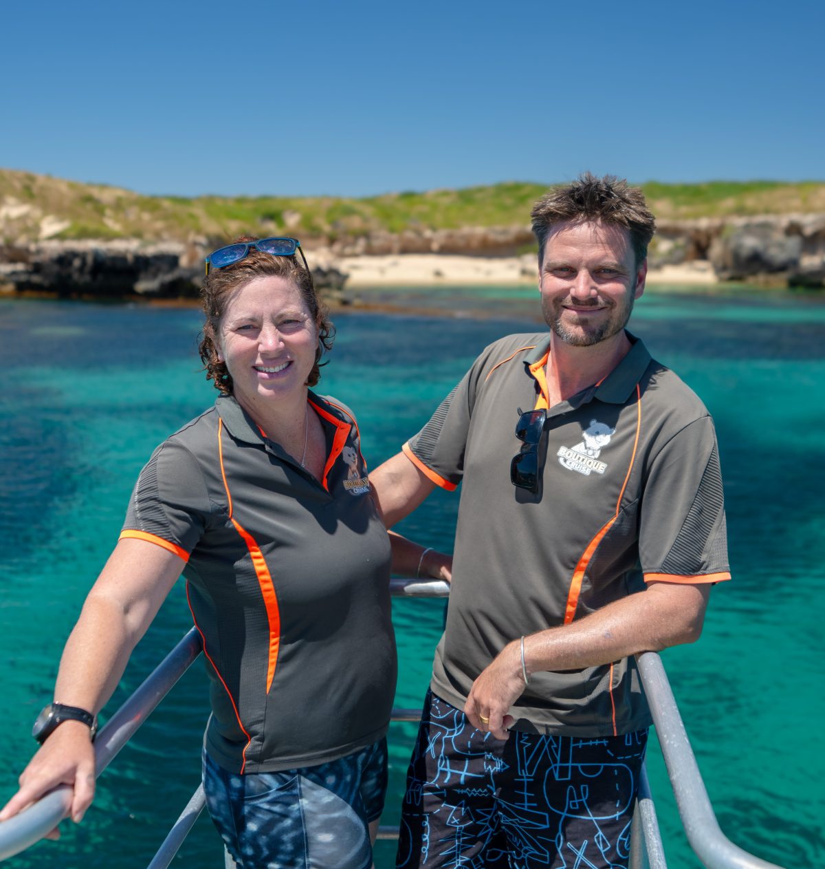Two people smiling on a boat deck with clear blue water and rocky coastline in the background.