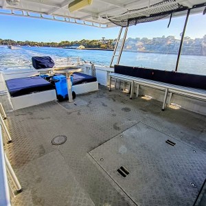 Interior of a boat with benches, table, and blue cushions, overlooking a calm blue lake and distant houses.