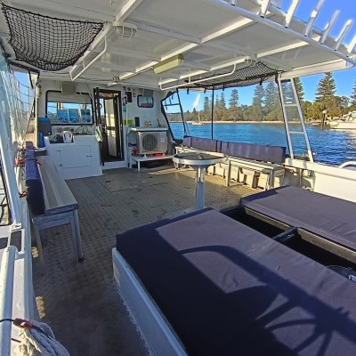 Interior of a boat with seating and tables, cruising on a sunny day with a view of the shore and trees.